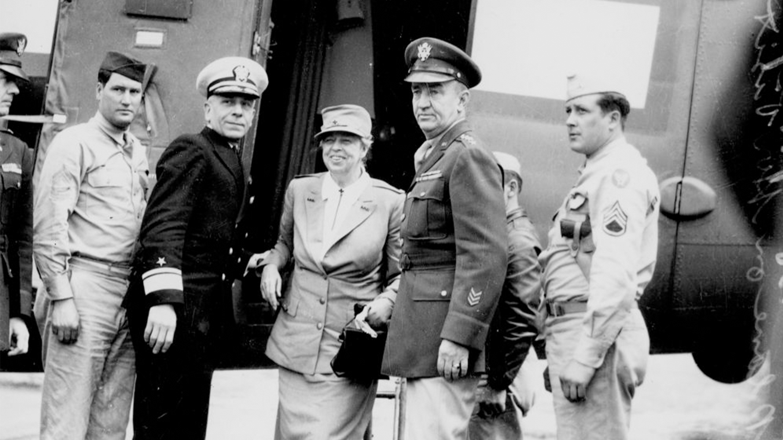 Eleanor Roosevelt disembarking from her flight in Brisbane. She is smiling and flanked by four military representatives.