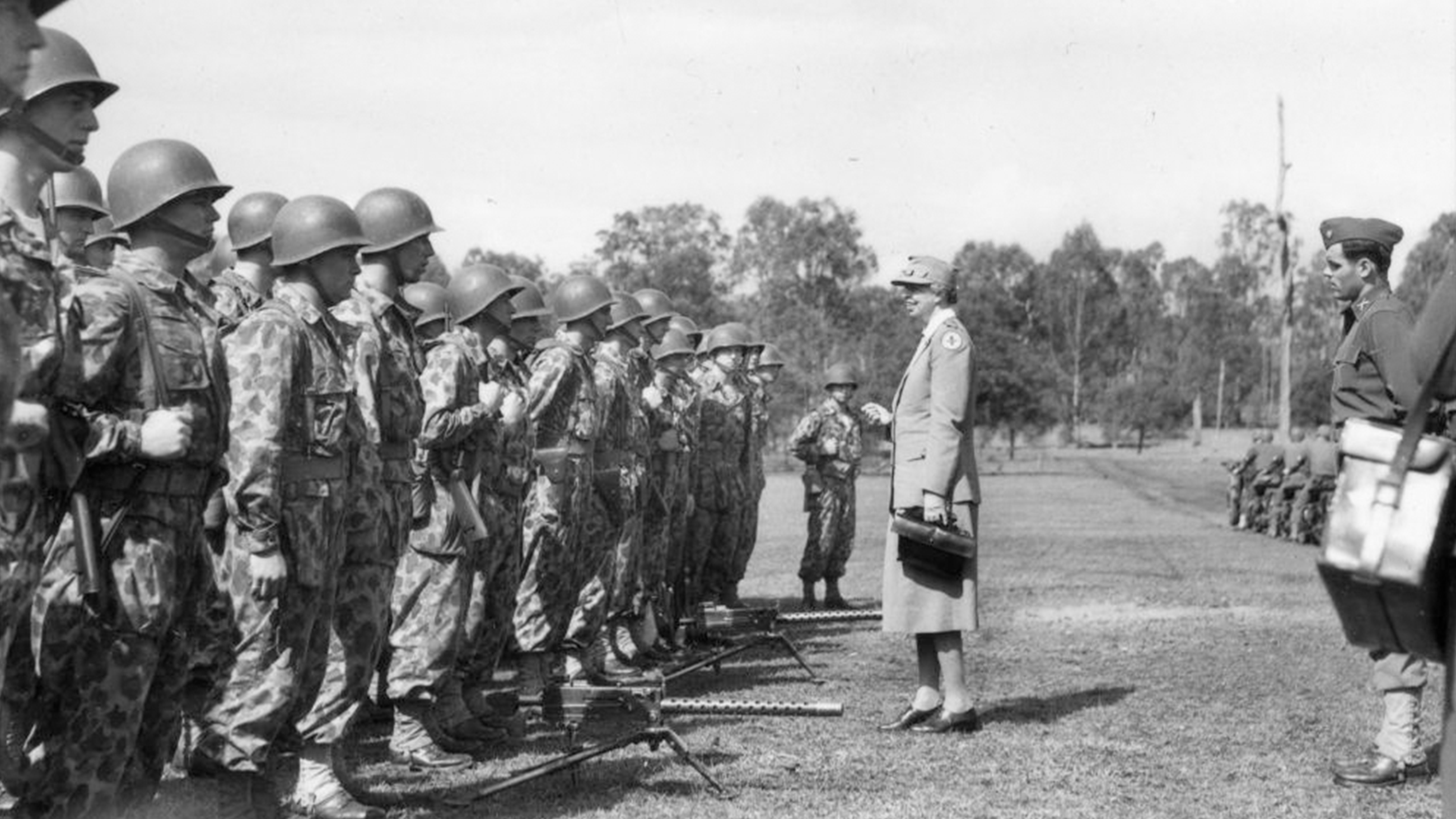 Eleanor Roosevelt stands in front of a line-up of soldiers in an open, outdoor clearing lined with trees. Eleanor is wearing her Red Cross uniform and smiling as she addresses the soldiers.