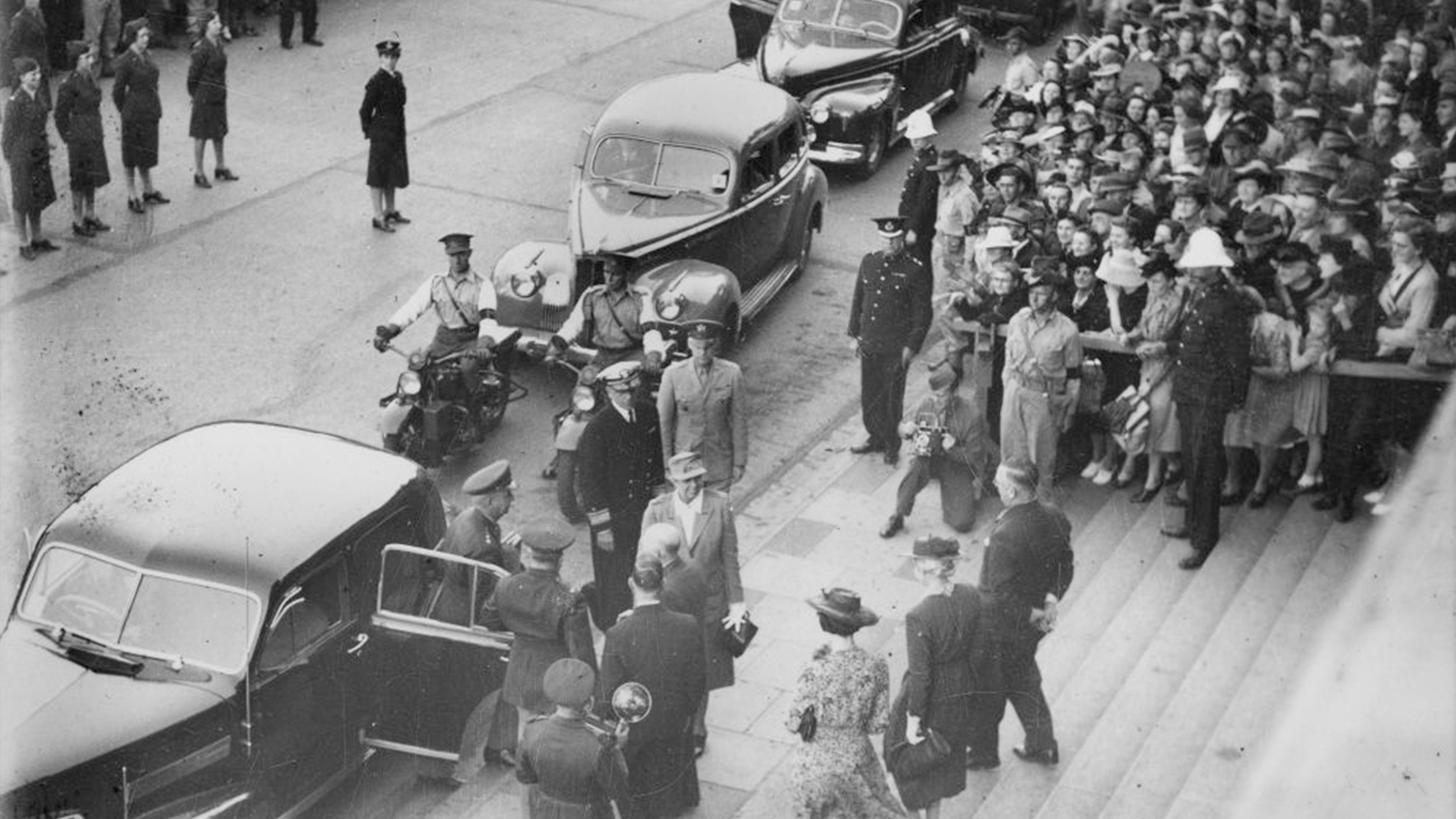 Eleanor Roosevelt stands on the steps of Brisbane City Hall and is greeted by government officials, press and excited members of the public. Her motorcade stretches back into the distance.