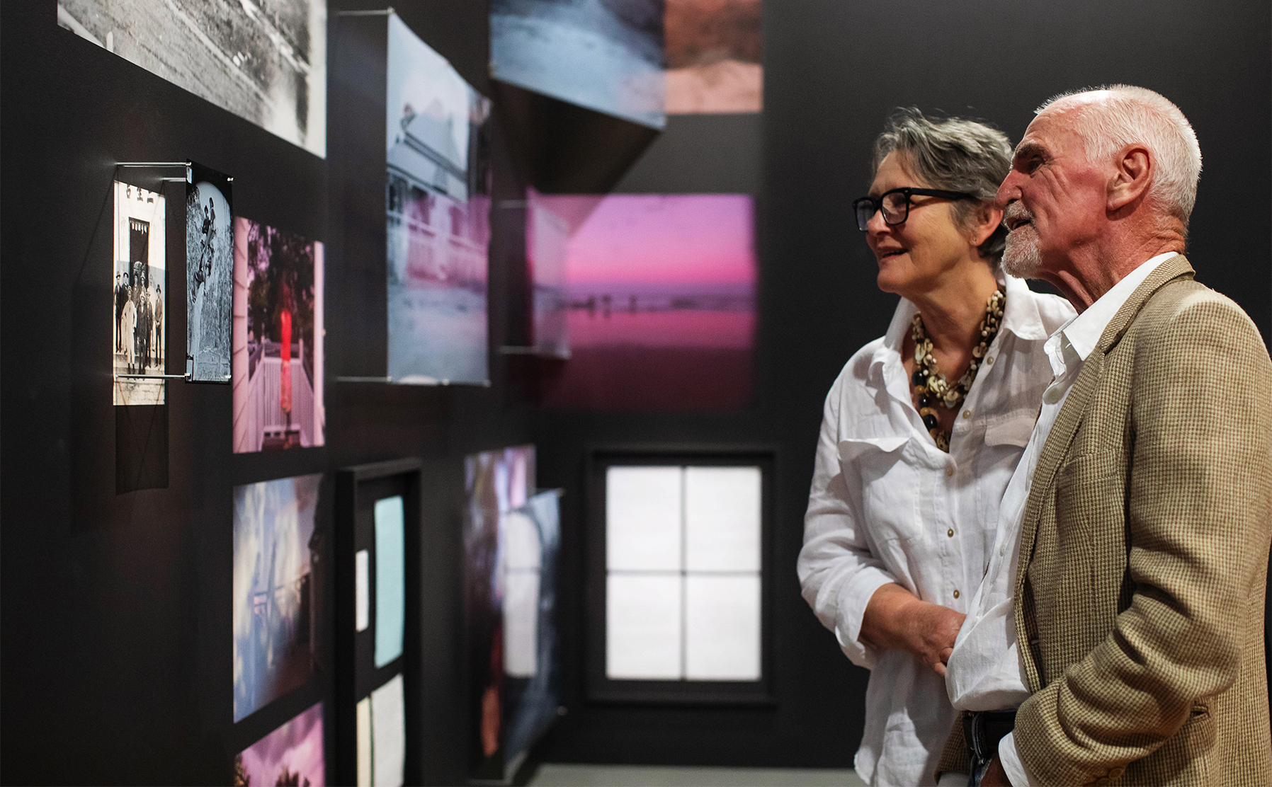 A couple smiling at a group of photos on a wall.