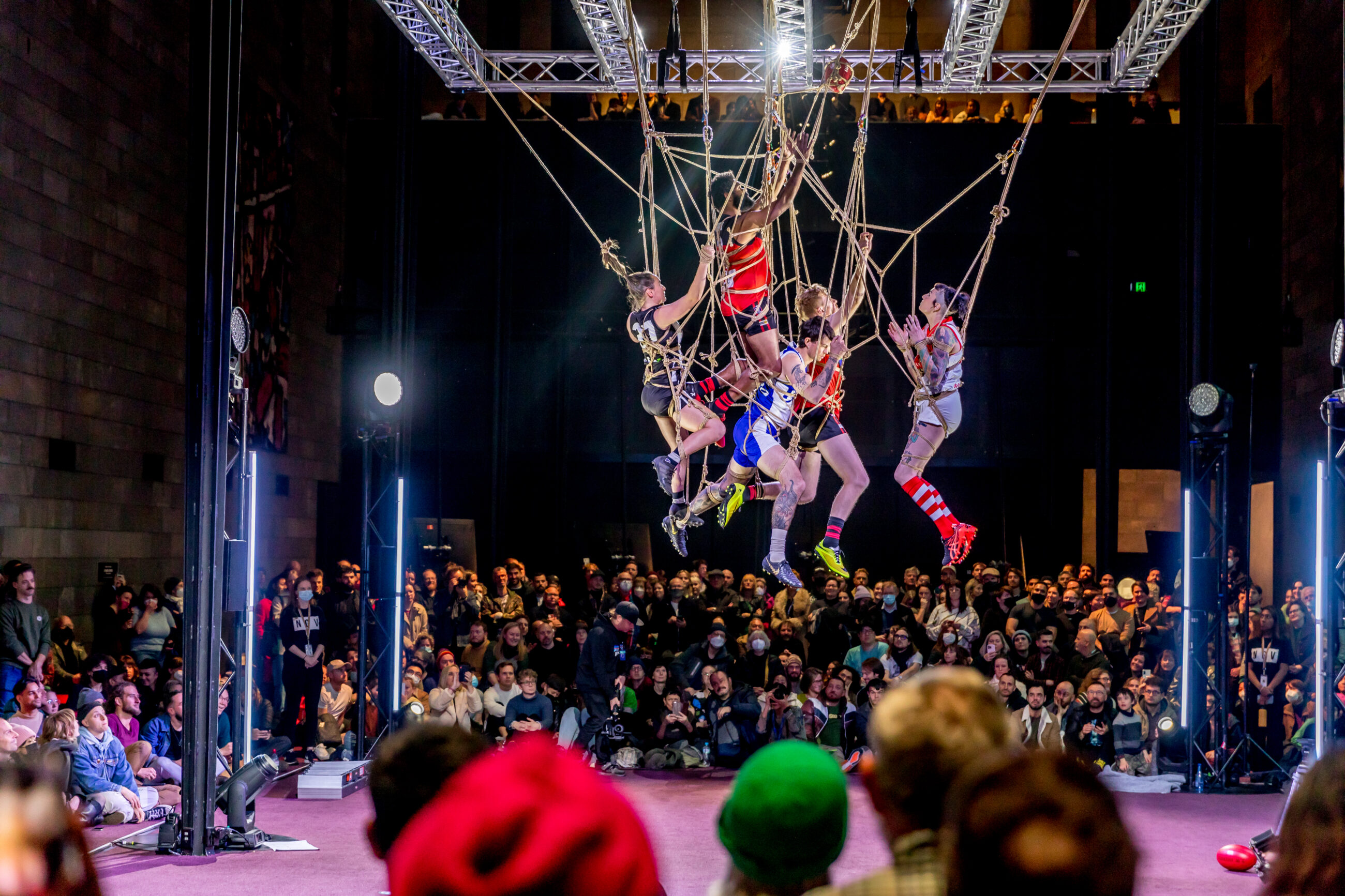 An array of people mixed within rope hanging from the ceiling with an audience watching